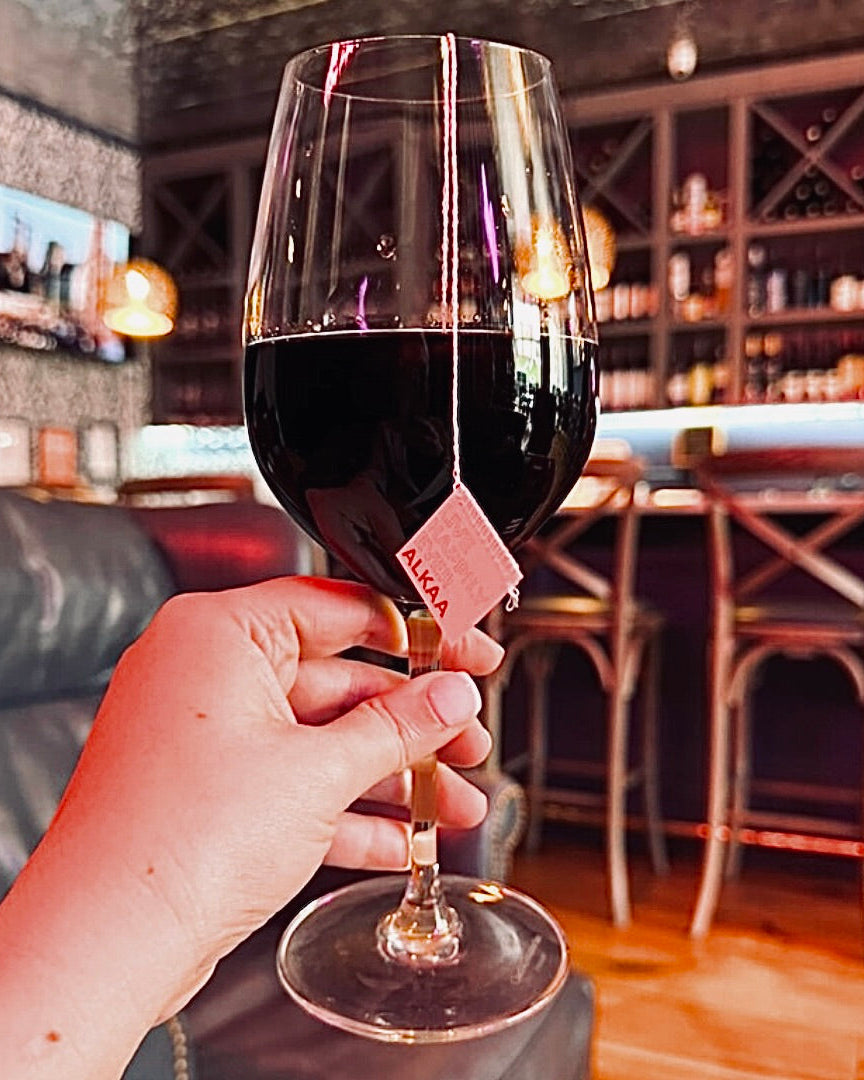Hand holding a glass of red wine containing an ALKAA sachet with a blurred bar background