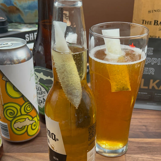 A open beer bottle with a Skinny style ALKAA sachet in the neck of the bottle sitting alongside a tall glass of beer that also contains a Skinny Sachet sitting on a wood surface.