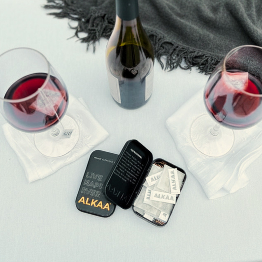 Two glasses of red wine, a bottle, and small containers labeled 'ALKAA' on a light surface with a dark cloth in the background.