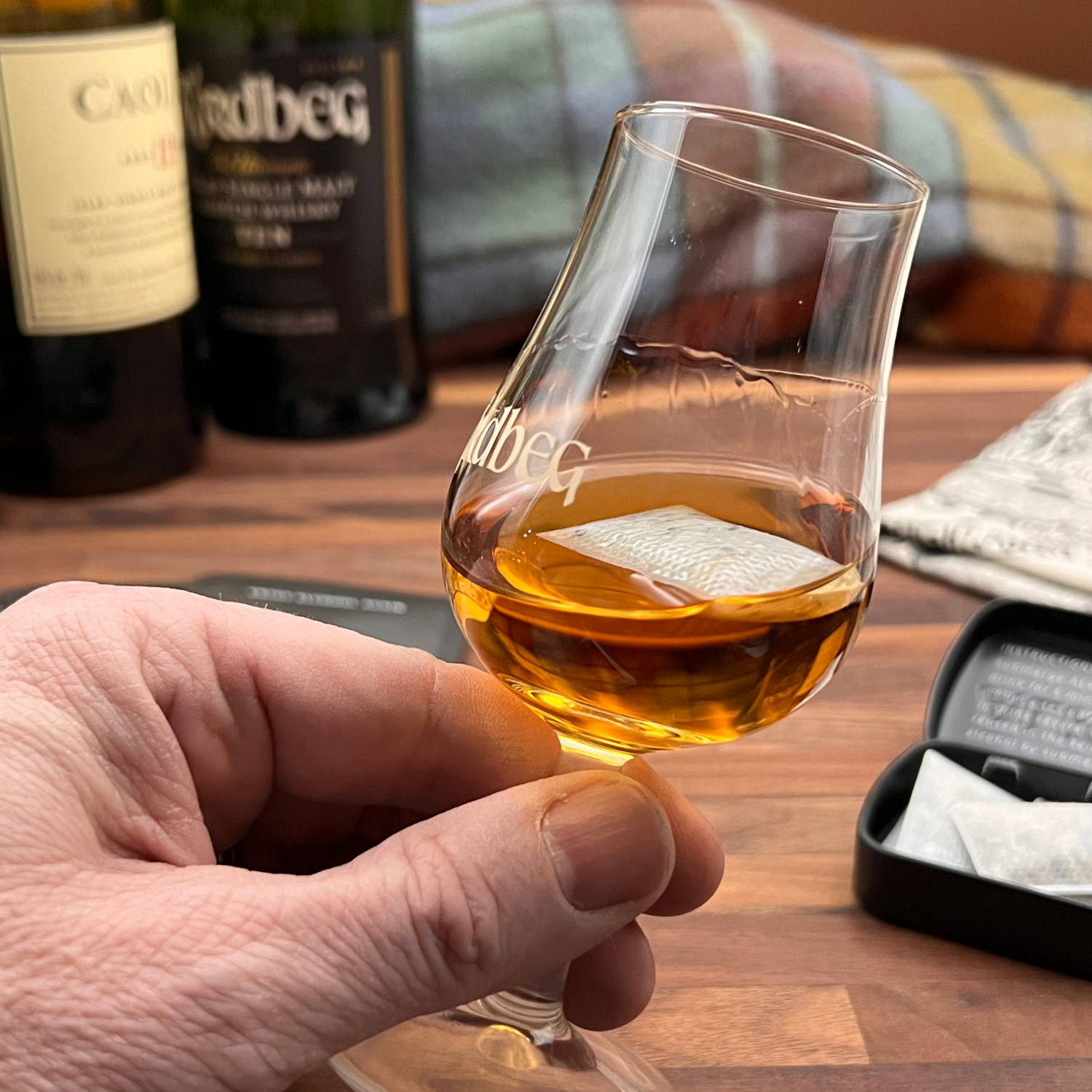 Hand holding a whisky glass containing the Dram style ALKAA sachet with bottles and a book in the background