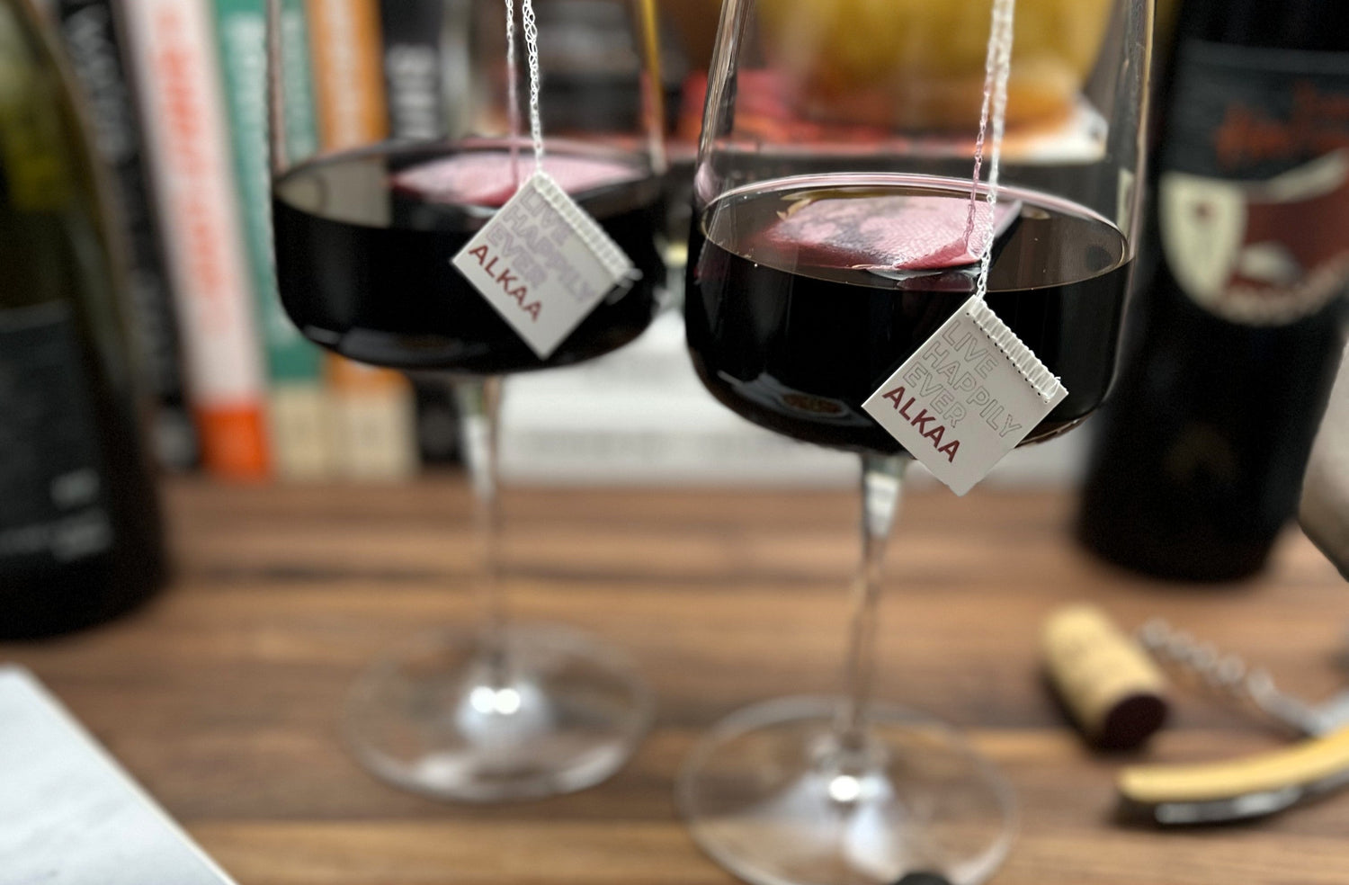 Two wine glasses with tea bag style Tagged ALKAA sachets labeled on a wooden table.