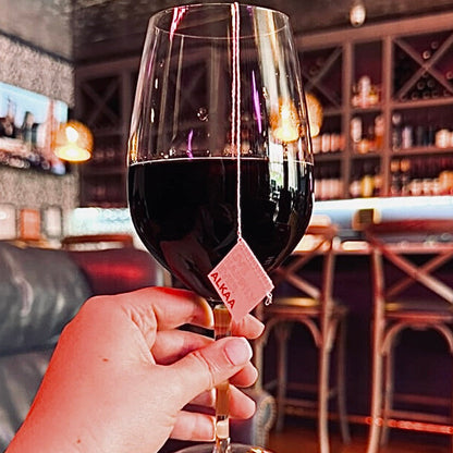 Hand holding a glass of red wine containing an ALKAA sachet with a blurred bar background