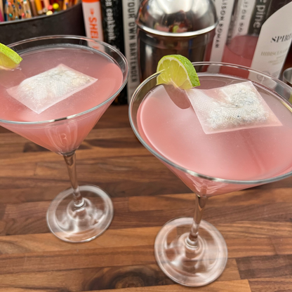 Two martini glasses filled with a pink liquid, garnished with a slice of lime and a floating ALKAA sachet, on a wooden surface with various bottles in the background.