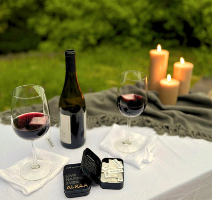 Wine bottle and glasses with ALKAA sachets and with candles on a picnic blanket outdoors. An open tin of ALKAA is in the foreground.