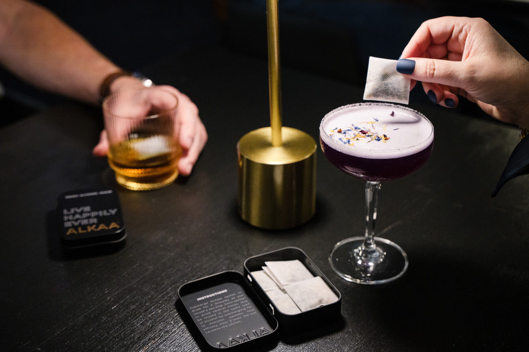 Person adding an alcohol filter to a drink on a dark surface with a tin of filters open and other items.