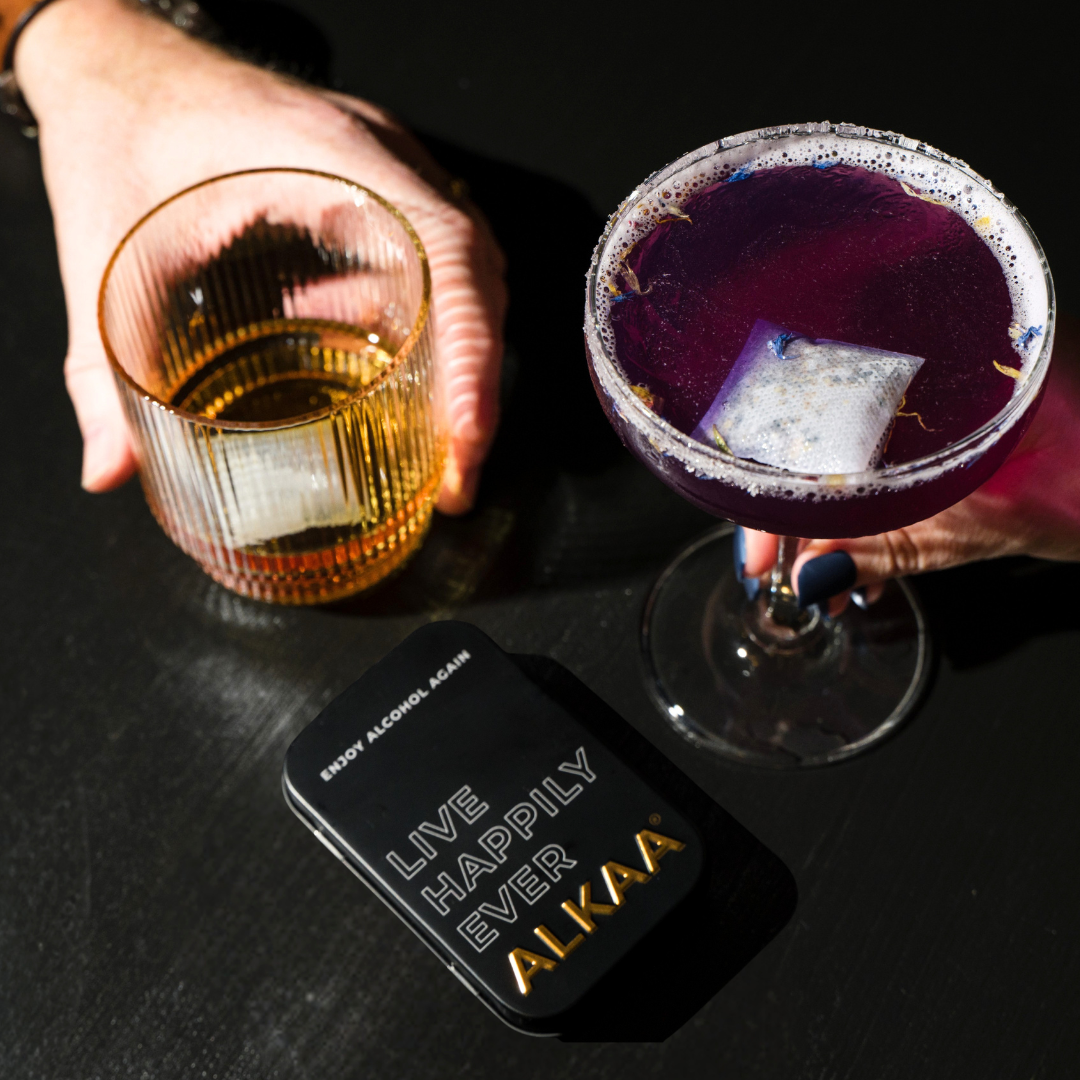 2 hands holding alcoholic beverages on a black background showing ALKAA tin and two alcohol purifying sachets.