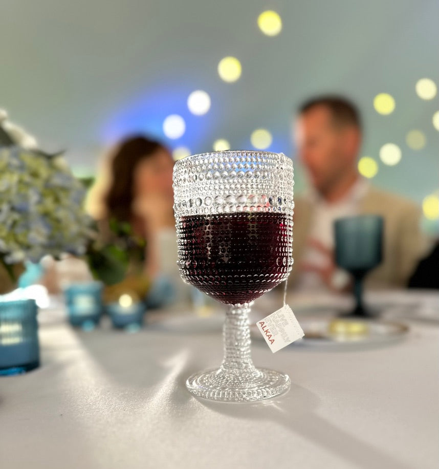 Wedding reception with two people talking in the background. A glass of red wine with an ALKAA sachet in the foreground on a white tablecloth.