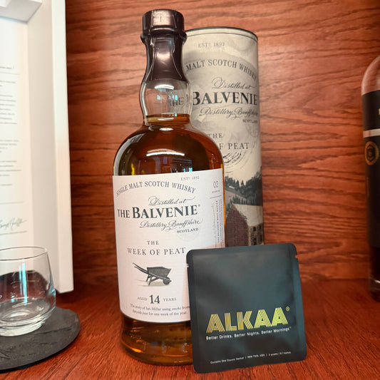 A bottle of Balvenie single malt Scotch whisky stands on a wooden shelf alongside its cylindrical tin packaging, a small black pouch labeled ALKAA, and a clear glass on a dark coaster.