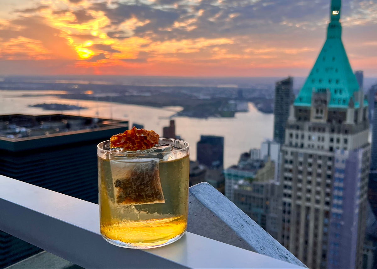 A cocktail with an ALKAA sachet rests on the ledge of a rooftop bar in Manhatten, NY. The setting sun in the background lights up the cocktail in the foreground. 