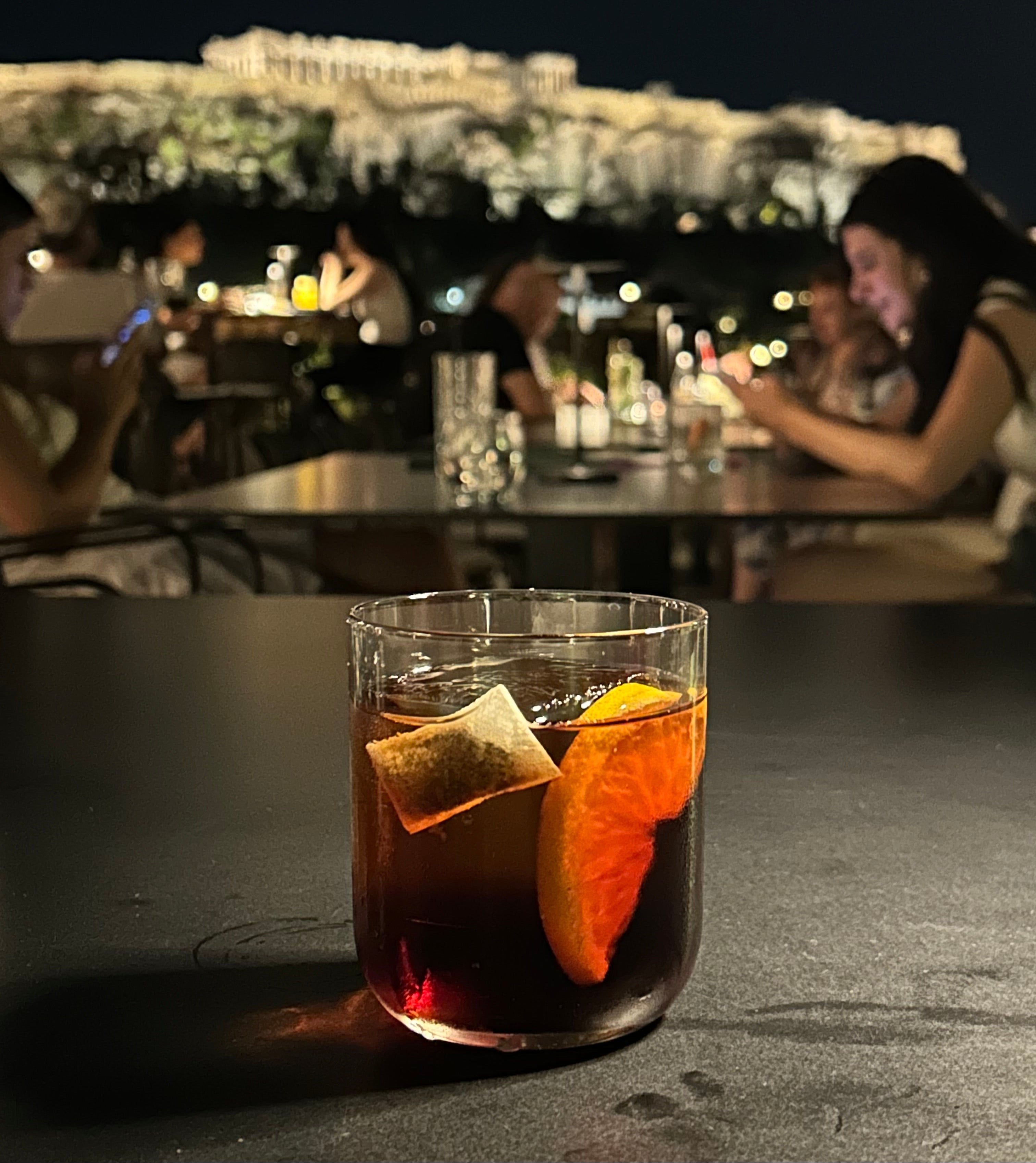 Close-up of a cocktail with an orange slice and an ALKAA sachet, blurred bar scene in the background