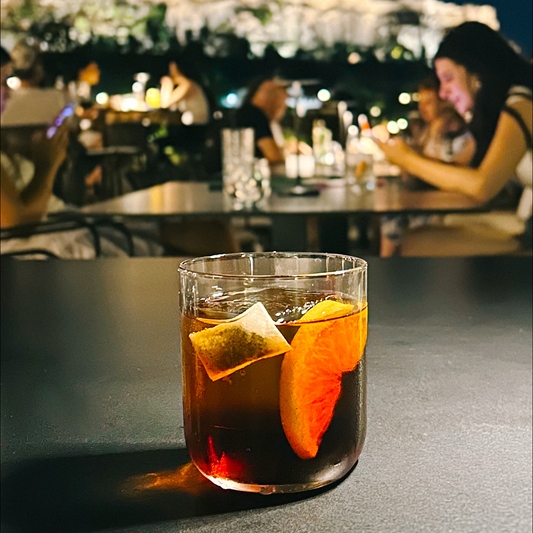 Cocktail with Dram sachet and orange slices on a table with a blurred background of people and lights.