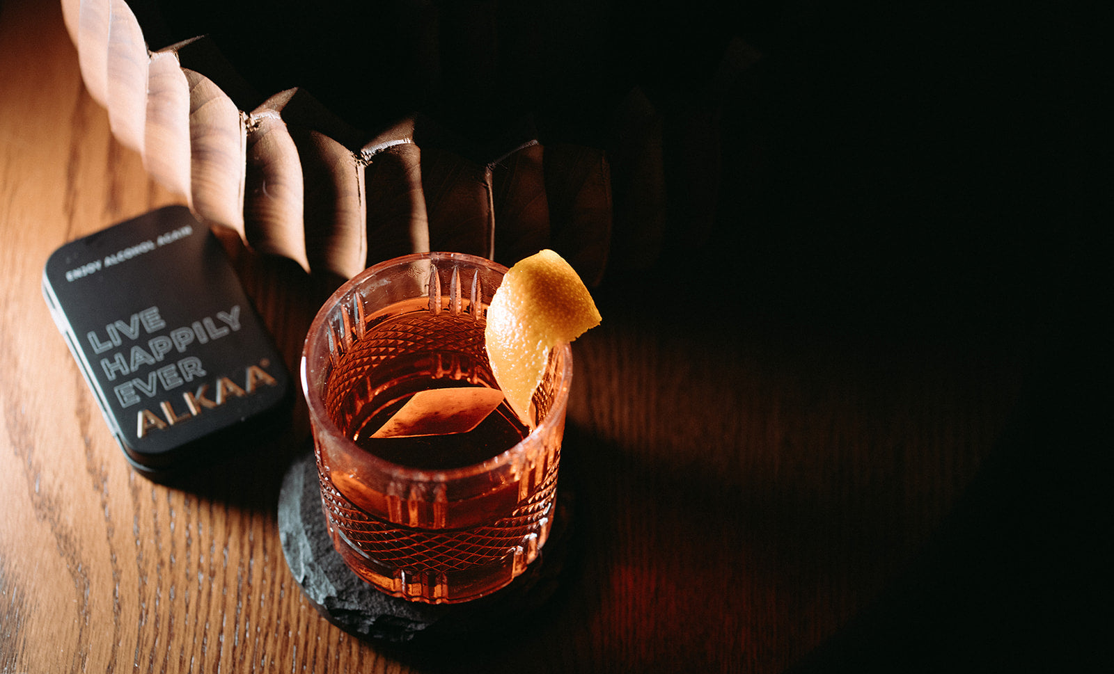 A textured clear glass tumbler filled with an amber-brown drink and garnished with a curled orange peel, resting on a dark coaster on a wooden table. A black tin labeled 'LIVE HAPPILY EVER AIKAA' sits beside it, with a blurred background and dramatic low lighting.