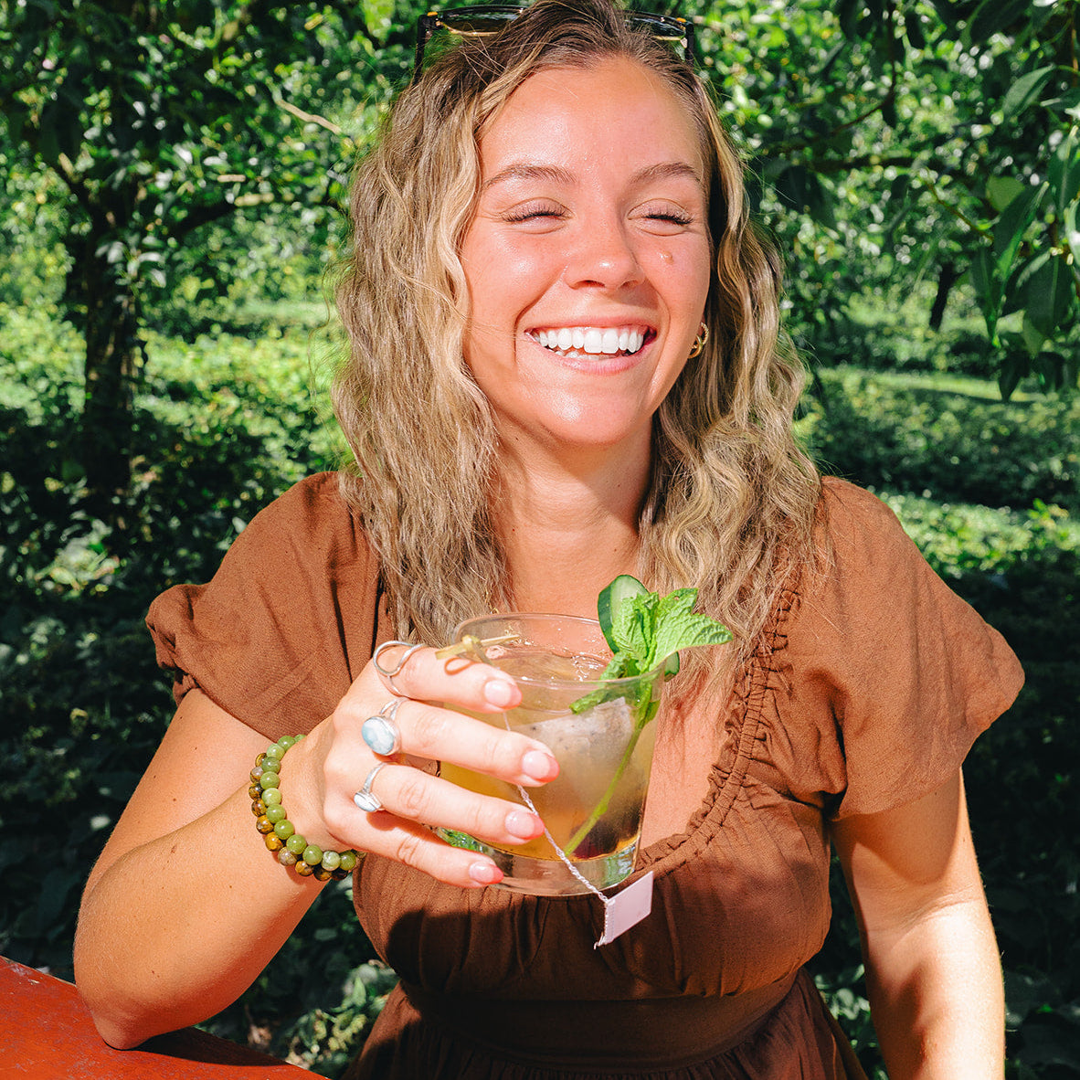 Woman in a brown dress holding a drink with a tagged ALKAA sachet outdoors with greenery in the background