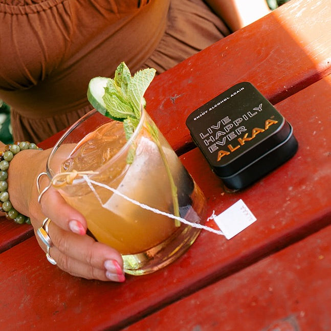 Hand holding a glass of tea with a lemon wedge and mint leaf, next to a black container labeled 'Alkaa' on a red surface.